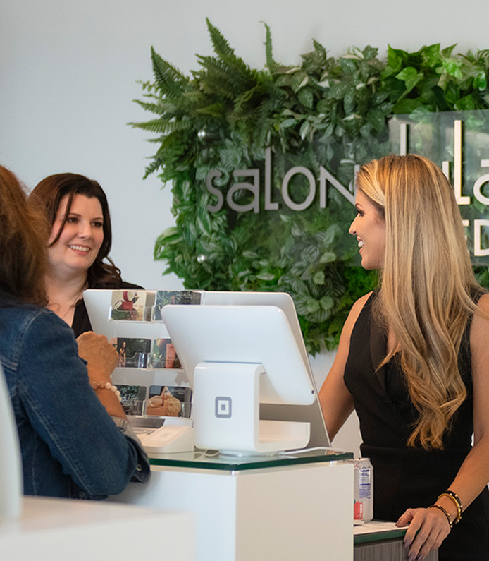 Two women stand at a salon reception desk, interacting with a receptionist, with a green plant wall and "salon" sign in the background. - Heart of Hospitality by Laura Dulay